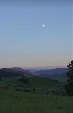 the moon is setting in the sky over a grassy field with mountains and trees behind it