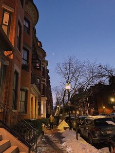 people are walking down the sidewalk in front of some buildings at night with snow on the ground