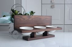 three white bowls sit on a wooden stand in front of a vase with green plants