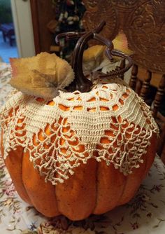 an orange pumpkin with crocheted doily sits on a floral tablecloth in front of a wooden chair