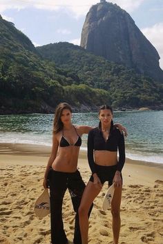 two women in bikinis standing on the beach with mountains in the background and water behind them