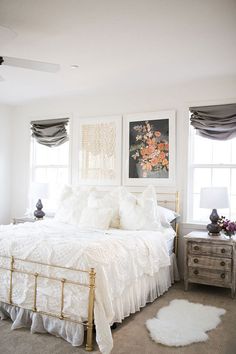 a bedroom with white bedding and pictures on the wall above it, along with two windows