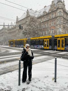 a woman standing in the snow next to a street with two yellow trains on it