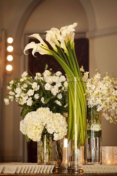 white flowers in vases sitting on a table with candles and napkins next to them