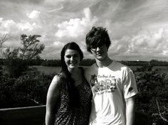 two people standing next to each other in front of some trees and water with clouds