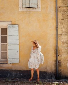 a woman standing in front of a yellow building wearing a white dress and hat with her hands on her head