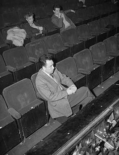 black and white photograph of man sitting in an empty auditorium with other people behind him