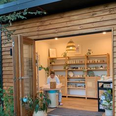 a woman standing in the doorway of a wooden building with shelves and pots on it