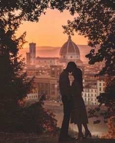 a man and woman standing next to each other in front of trees with the sun setting behind them