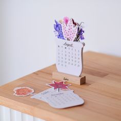 a wooden table topped with a calendar and flower vase filled with paper cutout flowers