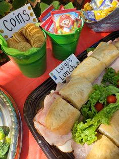 a table topped with sandwiches and salads next to plastic containers filled with chips, crackers and veggies