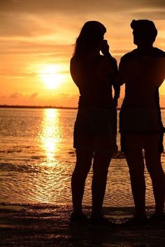 two people standing in front of the ocean watching the sun go down over the water