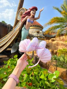 a hand is holding a pink flower in front of a fake mermaid and pirate ship