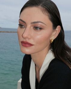 a woman with long hair wearing gold earrings and a black sweater by the water looking at the camera