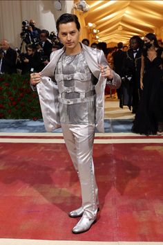 a man in silver suit and white shoes standing on red carpet with cameras around him