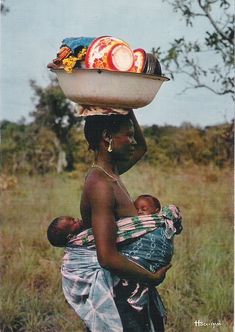 a woman carrying a baby in her arms with fruit on it's head,