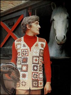 a man standing in front of a horse wearing a sweater and hat with his hand on his hip