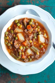 a white bowl filled with stew and vegetables on top of a blue tablecloth next to a spoon