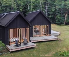 two black cabins in the woods with decking and chairs on it's sides