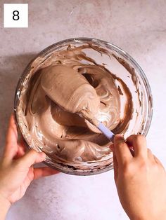 two hands holding a spoon over a bowl with chocolate frosting in it on a table