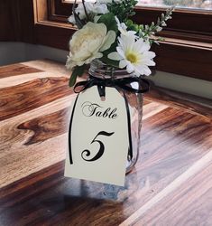 a table number sign with white flowers in a mason jar on a wood floor next to a window