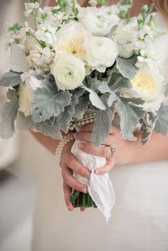 a bride holding a bouquet of white flowers and greenery in her hands, while wearing a wedding ring