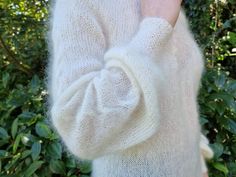 a woman is wearing a white sweater and holding her hand up to her face while standing in front of some bushes