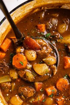 a pot filled with stew and carrots on top of a white countertop next to a wooden spoon