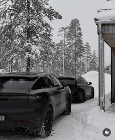 two cars are parked in the snow near a covered area with trees, and one is black