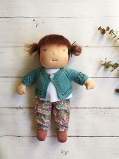 a doll with red hair wearing floral pants and a green cardigan is standing on a white wooden surface