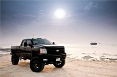 a black truck parked on top of a sandy beach next to the ocean with a boat in the background