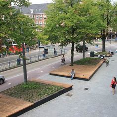 people are walking and sitting on benches in the middle of a city street with trees