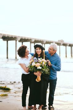 two women and an older man standing on the beach with flowers in front of them