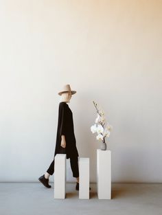 a woman in a hat is walking past some white blocks and a vase with flowers