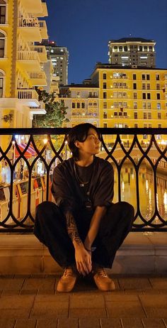 a man sitting on the ground in front of a city at night with buildings behind him