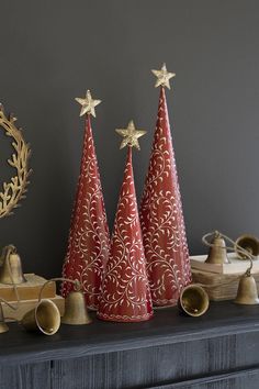 three red christmas trees on top of a mantle next to bells and other decorations in front of a gray wall