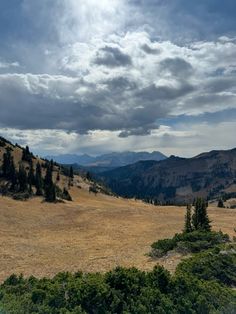 an open field with mountains in the distance and trees on the other side, under a cloudy sky