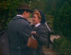 a man and woman standing next to each other in front of some green trees with a horse behind them