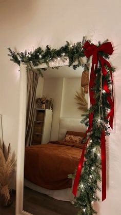 a mirror with a red bow on it next to a bed and christmas tree branches