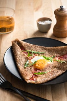 a black plate topped with an egg and bacon pita bread next to a glass of tea