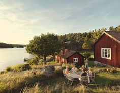 a group of people sitting around a table in front of a red house by the water