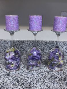 three wine glasses filled with purple flowers on top of a granite counter next to candles