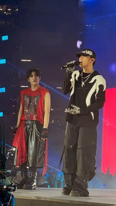 two people standing on stage with microphones in their hands and one person wearing a hat