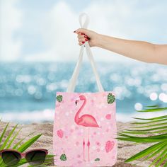 a pink flamingo shopping bag sitting on the beach with sunglasses and palm leaves around it