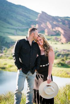 a man and woman standing next to each other in the grass with mountains behind them