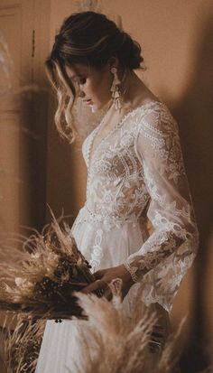 a woman in a white dress standing next to some dry grass and looking down at her bouquet