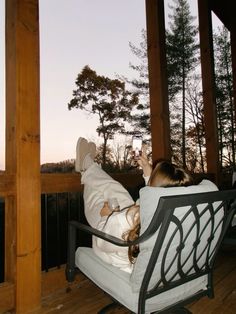 a woman laying on top of a white chair next to a wooden porch with trees in the background