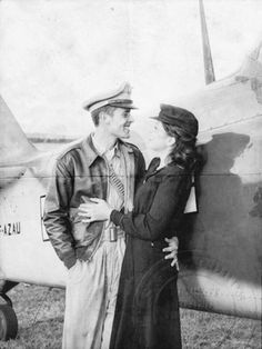 a man and woman standing in front of an old fashioned airplane with the pilot leaning against the wing