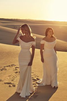 two beautiful women in white dresses walking through the sand dunes with footprints on the ground