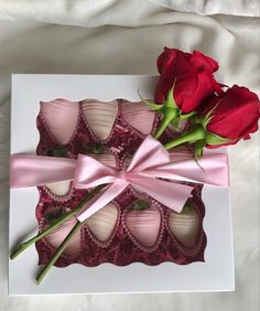 two red roses in a box with pink ribbon and chocolates wrapped in paper on a white sheet